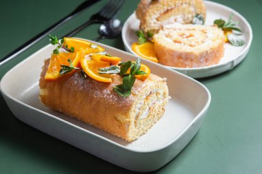 Parlak bir arka planda beyaz bir tabakta kremalı ve naneli turuncu sünger rulosu.