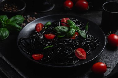Vişneli siyah spagetti ve siyah arka planda siyah bir tabakta fesleğen..