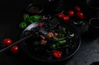 Ton balıklı siyah spagetti, vişneli domates ve siyah bir tabakta fesleğen..