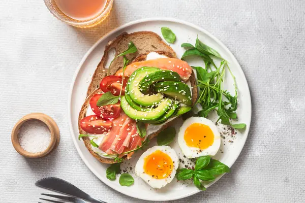  Somonlu sandviç, avokado, domates ve yumurta yakın plandaki beyaz tabakta. Sağlıklı kahvaltı.
