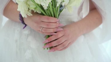 Yakın plan çekimde gelinlerin elleri pembe manikürlü, beyaz bir gelinlikle süslenirken bir buket beyaz çiçeği kucaklıyor. Düğün ve gelinlik için ideal içerik