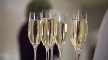 Resmi giyinmiş bir garson zarif bir şekilde sofistike bir etkinlikte gümüş tepside dört bardak şampanya servis ediyor. Bu görkemli ortamda içkilere uzanan eller görülüyor.