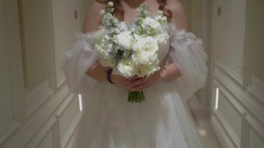 Zarif gelin, omuz hizasında olmayan bir elbise içinde, zarif bir çiçek buketini zarif bir şekilde güzel dekore edilmiş bir koridorda, zamansız romantizm ve onun özel düğün gününde gelinlik güzelliğinde tutuyor.