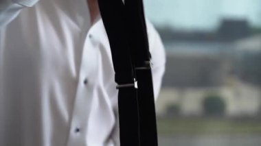 Modern bir ortamda, beyaz gömlekli bir adam siyah pantolon askılarını düzeltir, zarif ve profesyonel bir görünümü somutlaştırır, biraz stil ve keskinlik, gösterişli tımar ve çalışmaya hazır olma ile.