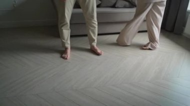 Ahşap zeminde insan ayağı. Genç kadın çıplak ayakla odada daireler çiziyor ve dans ediyor. Beyaz pantolonlu bir kadın. Tanımlanamayan kız ve erkek bacakları. Evdeki çift..