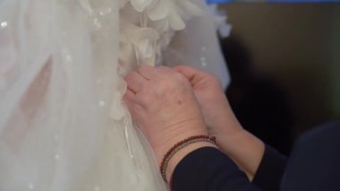 Ayrıntılı bir yakın çekim, zarif bir gelinliğin bağcıklarını nazikçe bağlayan, özel gün için titiz hazırlıklar yapan zarif güzellik ve aşkla dolu bir düğün törenini gözler önüne seriyor.