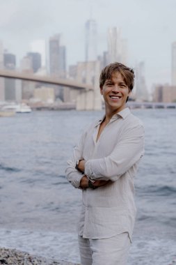 Neşeli genç bir adam, arka planda nefes kesen bir şehir silueti ile, ışıldayan suyun yanında poz verir. Brooklyn Köprüsü, New York 'ta Manhattan.