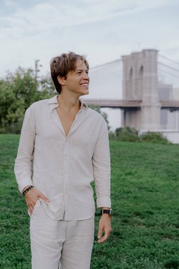 Tarz sahibi bir birey, arkasında Brooklyn Köprüsü olan canlı yeşil bir parkta güvenle duruyor.