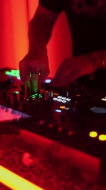 Canlı bir gece kulübünde renkli ışıklandırmayla müziği karıştıran bir DJ 'in yakın çekimleri. DJ 'in elleri çeşitli kontrolleri ayarlıyor ve enerjik bir atmosfer yaratıyor..