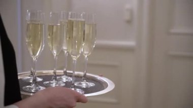Resmi giyinmiş bir garson zarif bir şekilde sofistike bir etkinlikte gümüş tepside dört bardak şampanya servis ediyor. Bu görkemli ortamda içkilere uzanan eller görülüyor.
