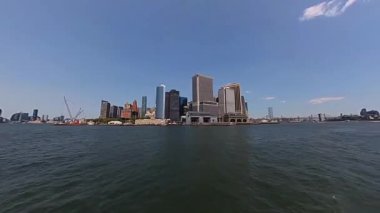 İkonik New York siluetinin berrak gökyüzüne ve sakin bir su ön planına karşı çarpıcı panoramik görüntüsü.
