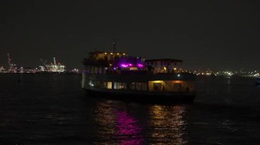 Canlı ve canlı bir parti teknesi gece boyunca yüzüyor, suyu aydınlatıyor ve çarpıcı şehrin ufuk çizgisini aydınlatıyor