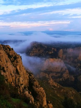 A dramatic mountain landscape shrouded in fog, with cliffs rising above a valley immersed in mist. The sky at dusk offers warm tones that contrast with the cold atmosphere of the fog clipart