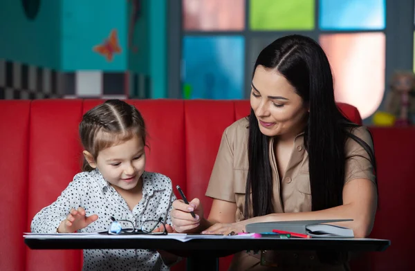 Mother helping her daughter with homework. Portrait of a smiling young mother and her little daughter drawing together in cafe. Happy childhood