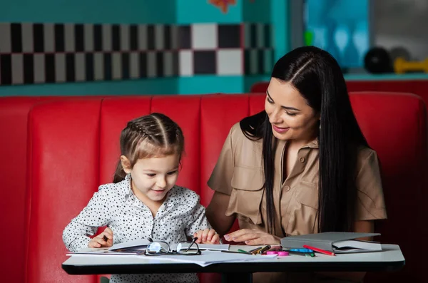 Mother helping her daughter with homework. Portrait of a smiling young mother and her little daughter drawing together in cafe. Happy childhood