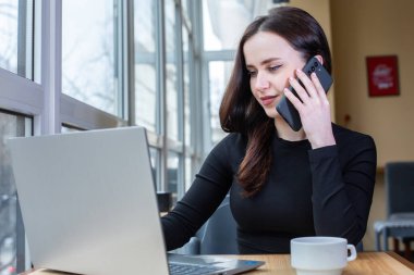 Portrait of smiling young woman using mobile phone while sitting in in coffee shop. White female freelancer connecting to internet on mobile phone.