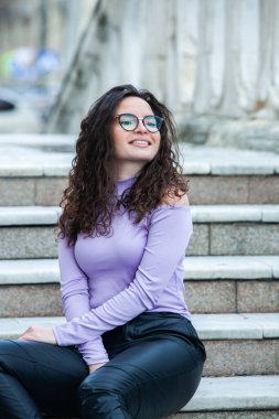 Esmer, kıvırcık saçlı, gözlüklü güzel bir kadın. Şehirde güneşin tadını çıkarıyor..