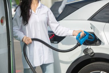 Genç bir iş kadını elektrikli arabasına EV şarj istasyonunda yakıt ikmali yapıyor. Çevre dostu araç kavramı. Elektrikli araba konsepti. Yeşil seyahat.