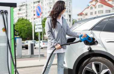 Genç bir iş kadını elektrikli arabasına EV şarj istasyonunda yakıt ikmali yapıyor. Çevre dostu araç kavramı. Elektrikli araba konsepti. Yeşil seyahat.