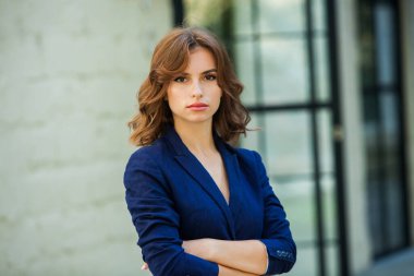 Uzun kahverengi saçlı, mavi ceketli, bir ofis binasının arka planında duran güzel bir genç kadının portresi. güçlü ve kendinden emin iş kadını