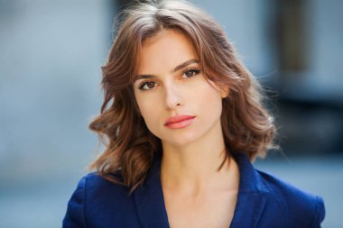 Uzun kahverengi saçlı, mavi ceketli, bir ofis binasının arka planında duran güzel bir genç kadının portresi. güçlü ve kendinden emin iş kadını