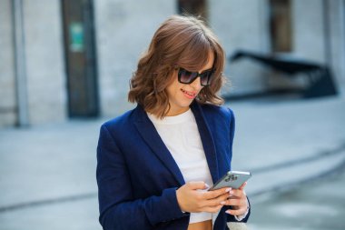 Uzun kahverengi saçlı, mavi ceketli, bir ofis binasının arka planında duran güzel bir genç kadının portresi. güçlü ve kendinden emin iş kadını