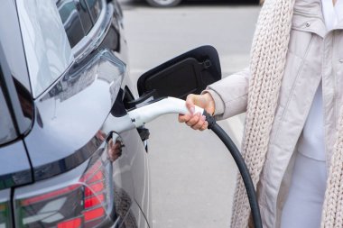 Şehirdeki bir şarj istasyonunda genç bir kadın elektrikli arabasını şarj ediyor. Eko yakıt konsepti. Çevre dostu ulaşım kavramı. Şarj istasyonundan şarj ediliyor..