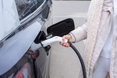 Şehirdeki bir şarj istasyonunda genç bir kadın elektrikli arabasını şarj ediyor. Eko yakıt konsepti. Çevre dostu ulaşım kavramı. Şarj istasyonundan şarj ediliyor..