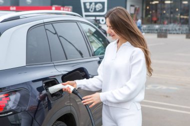 Şehirdeki bir şarj istasyonunda genç bir kadın elektrikli arabasını şarj ediyor. Eko yakıt konsepti. Çevre dostu ulaşım kavramı. Şarj istasyonundan şarj ediliyor..