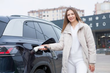 Şehirdeki bir şarj istasyonunda genç bir kadın elektrikli arabasını şarj ediyor. Eko yakıt konsepti. Çevre dostu ulaşım kavramı. Şarj istasyonundan şarj ediliyor..