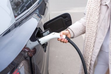 Şehirdeki bir şarj istasyonunda genç bir kadın elektrikli arabasını şarj ediyor. Eko yakıt konsepti. Çevre dostu ulaşım kavramı. Şarj istasyonundan şarj ediliyor..