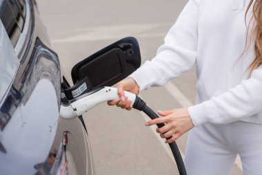 Şehirdeki bir şarj istasyonunda genç bir kadın elektrikli arabasını şarj ediyor. Eko yakıt konsepti. Çevre dostu ulaşım kavramı. Şarj istasyonundan şarj ediliyor..