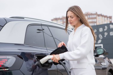 Şehirdeki bir şarj istasyonunda genç bir kadın elektrikli arabasını şarj ediyor. Eko yakıt konsepti. Çevre dostu ulaşım kavramı. Şarj istasyonundan şarj ediliyor..