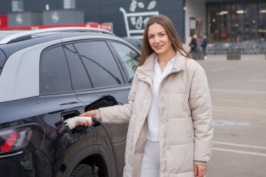 Şehirdeki bir şarj istasyonunda genç bir kadın elektrikli arabasını şarj ediyor. Eko yakıt konsepti. Çevre dostu ulaşım kavramı. Şarj istasyonundan şarj ediliyor..