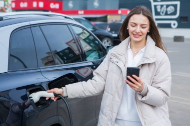 Şehirdeki bir şarj istasyonunda genç bir kadın elektrikli arabasını şarj ediyor. Eko yakıt konsepti. Çevre dostu ulaşım kavramı. Şarj istasyonundan şarj ediliyor..