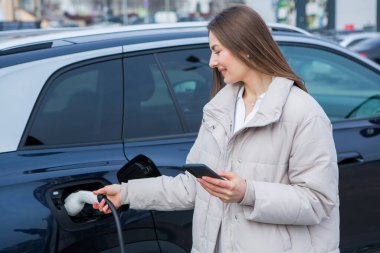 Şehirdeki bir şarj istasyonunda genç bir kadın elektrikli arabasını şarj ediyor. Eko yakıt konsepti. Çevre dostu ulaşım kavramı. Şarj istasyonundan şarj ediliyor..