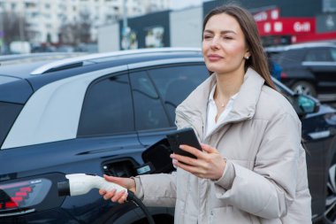 Şehirdeki bir şarj istasyonunda genç bir kadın elektrikli arabasını şarj ediyor. Eko yakıt konsepti. Çevre dostu ulaşım kavramı. Şarj istasyonundan şarj ediliyor..