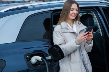 Şehirdeki bir şarj istasyonunda genç bir kadın elektrikli arabasını şarj ediyor. Eko yakıt konsepti. Çevre dostu ulaşım kavramı. Şarj istasyonundan şarj ediliyor..