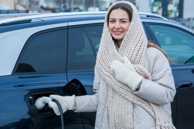 Şehirdeki bir şarj istasyonunda genç bir kadın elektrikli arabasını şarj ediyor. Eko yakıt konsepti. Çevre dostu ulaşım kavramı. Şarj istasyonundan şarj ediliyor..