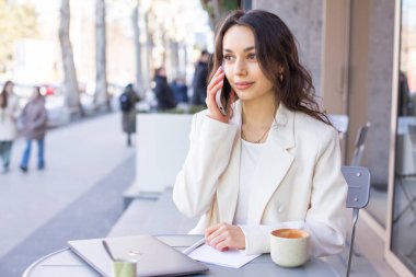 Zarif beyaz ceketli güzel bir bayan şehirdeki bir kafede telefonla konuşuyor.