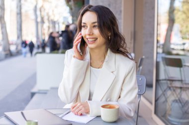 Zarif beyaz ceketli güzel bir bayan şehirdeki bir kafede telefonla konuşuyor.