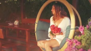 Güzel bir kadın elinde bir buket kırmızı gülle ahşap bir salıncakta oturur ve parlak bir ışık efektiyle.