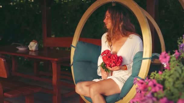美しい女性は手に赤いバラの花束を持って木製のスイングに座っている — ストック動画