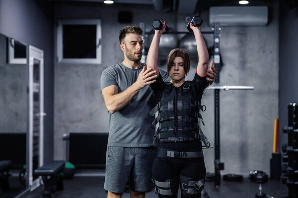 Female and male confident athletes train indoors at the gym. Woman is dressed in EMS technology in a gym and lifts dumbbells over her head while her trainer helps her perform a new exercise concept — Stock Image Female Male Confident Athletes Train Indoors Gym Woman Dressed Ems Royalty Free Stock Photos