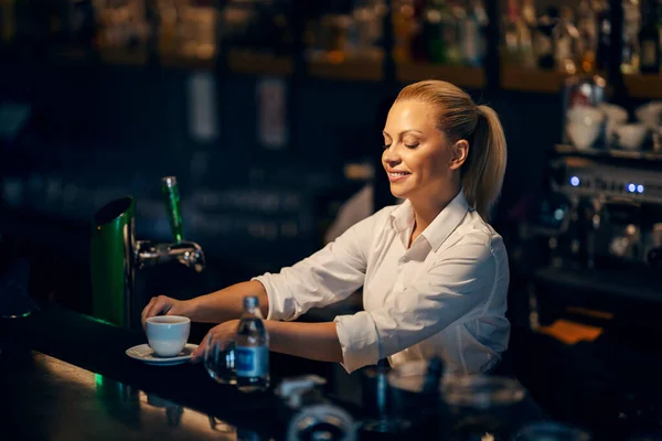 A happy female bartender is serving espresso while standing behind a bar counter in a bar.
