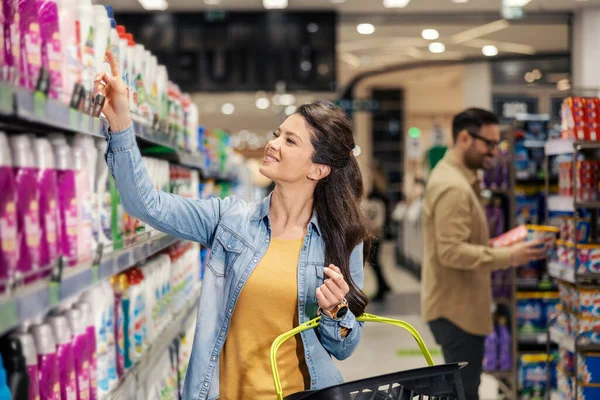 A smiling woman is taking detergent from shelf in hypermarket and buying it.