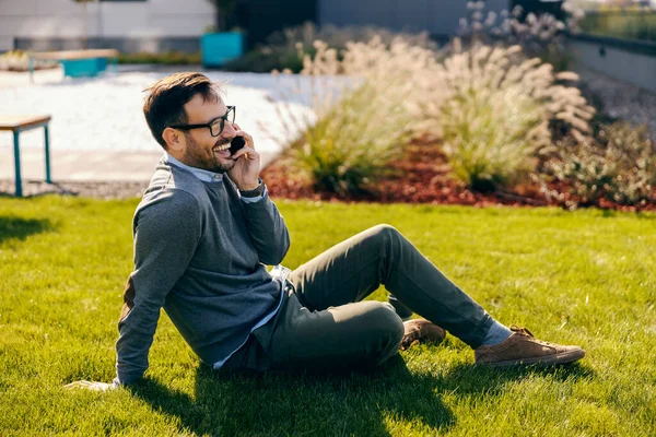 A trendy businessman smiles and talks on the phone while sits on a grass.