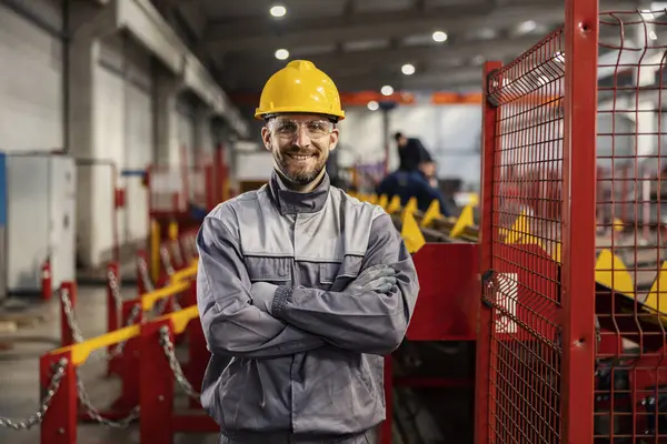 Proud metallurgy and heavy industry worker stands in a factory with arms crossed and smiles at the camera.