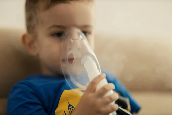 Portrait of an ill little boy inhaling himself at home with inhalator.