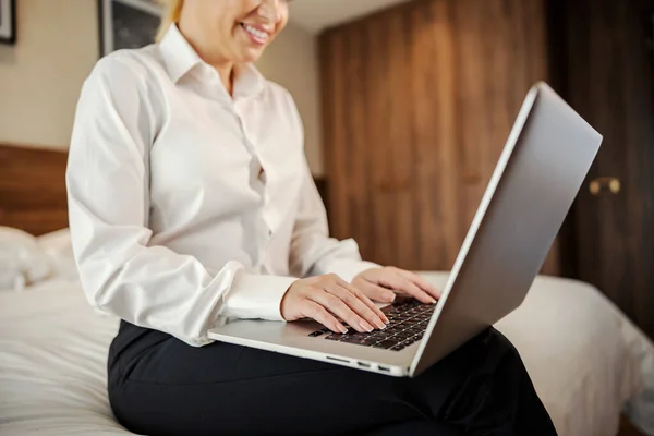 A happy businesswoman on a business trip is sitting in a hotel room and checking on her e-mail on a laptop.